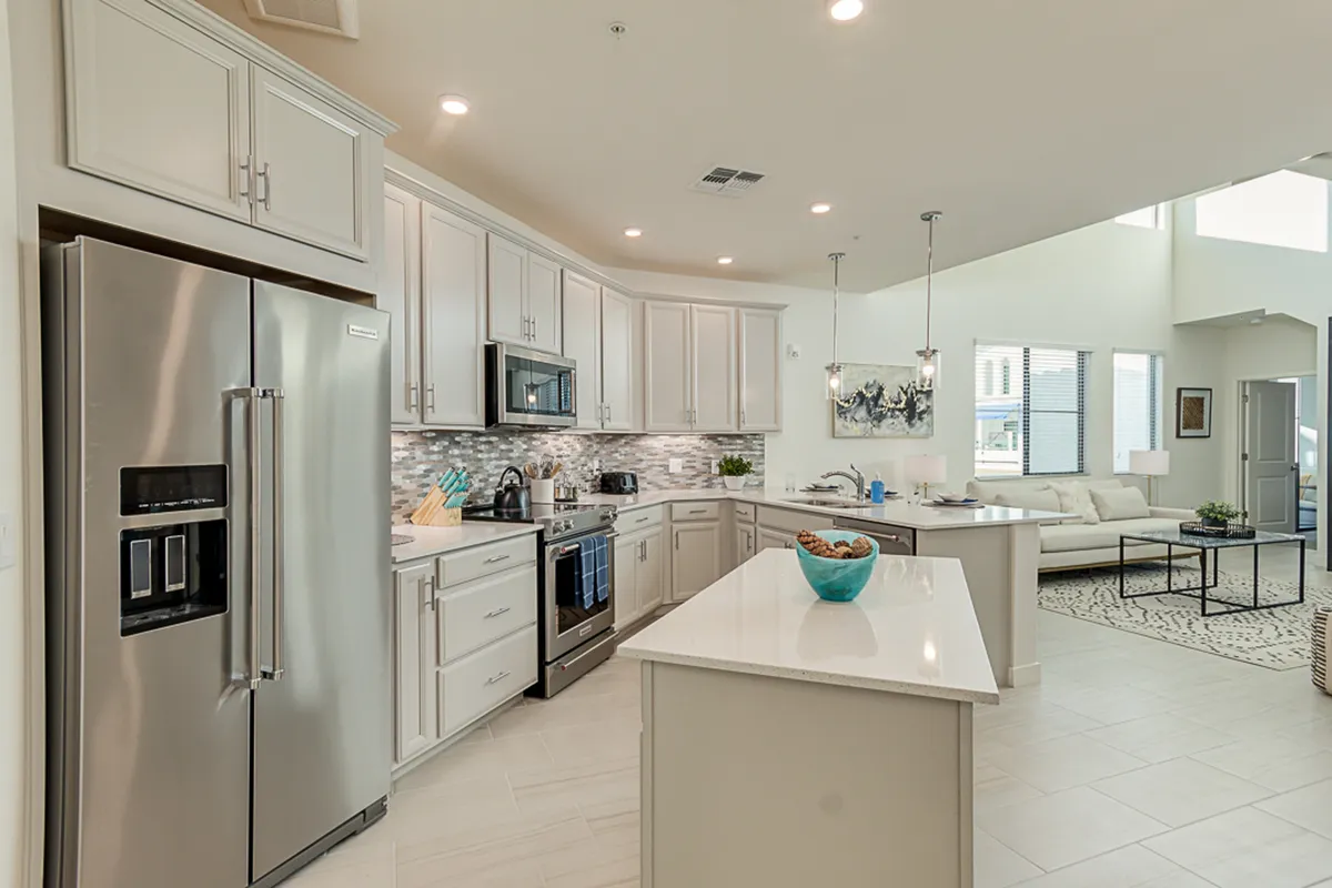A modern kitchen scene featuring a large island topped with a turquoise bowl, an impressive stainless steel refrigerator, contemporary light fixtures, and a view that extends into an open living area.