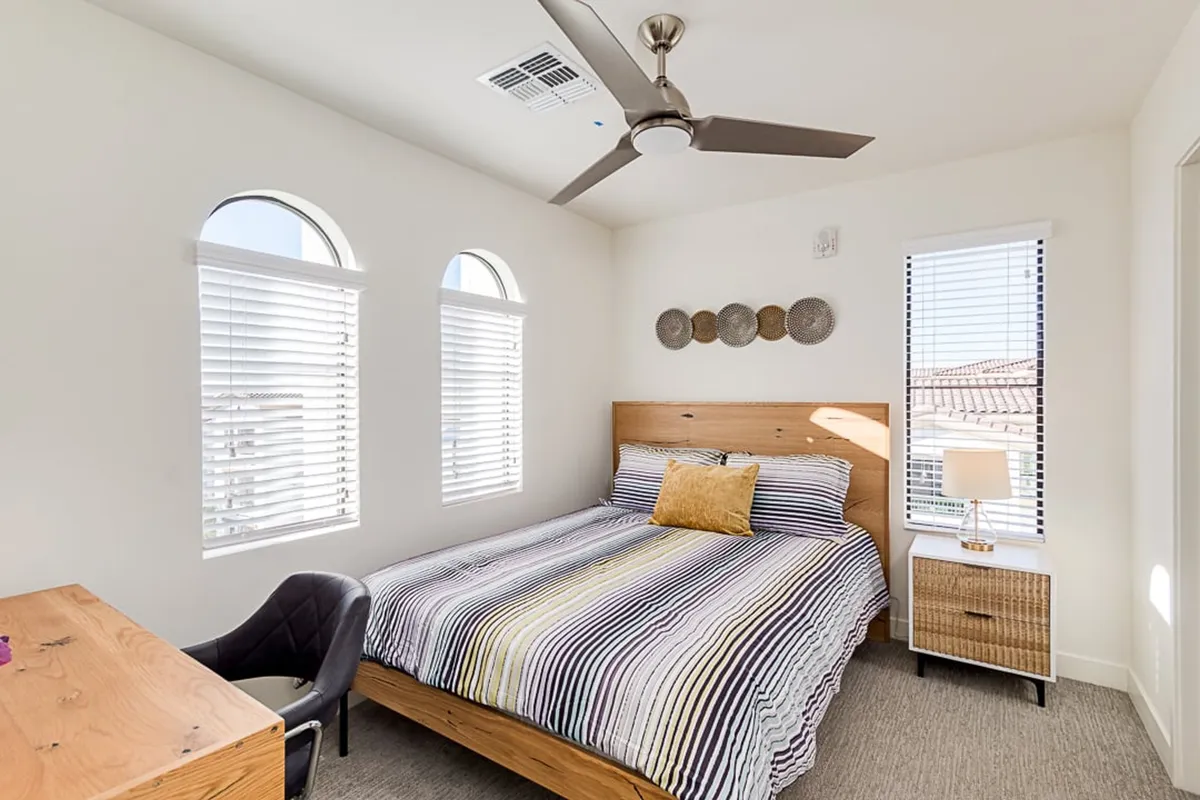A modern lofted bedroom/office with a bed featuring striped bedding, a wooden headboard, desk and chair, and a small bedside lamp. The workspace is neatly integrated into the sleeping area, providing a practical setup for work or study.