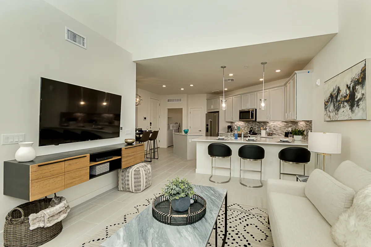 This image showcases a spacious, modern living area with a large TV as a focal point, a kitchen island with stylish seating, and decorative artwork on the walls. Natural light floods in, highlighting the clean lines and contemporary furnishings designed for both relaxation and socializing.