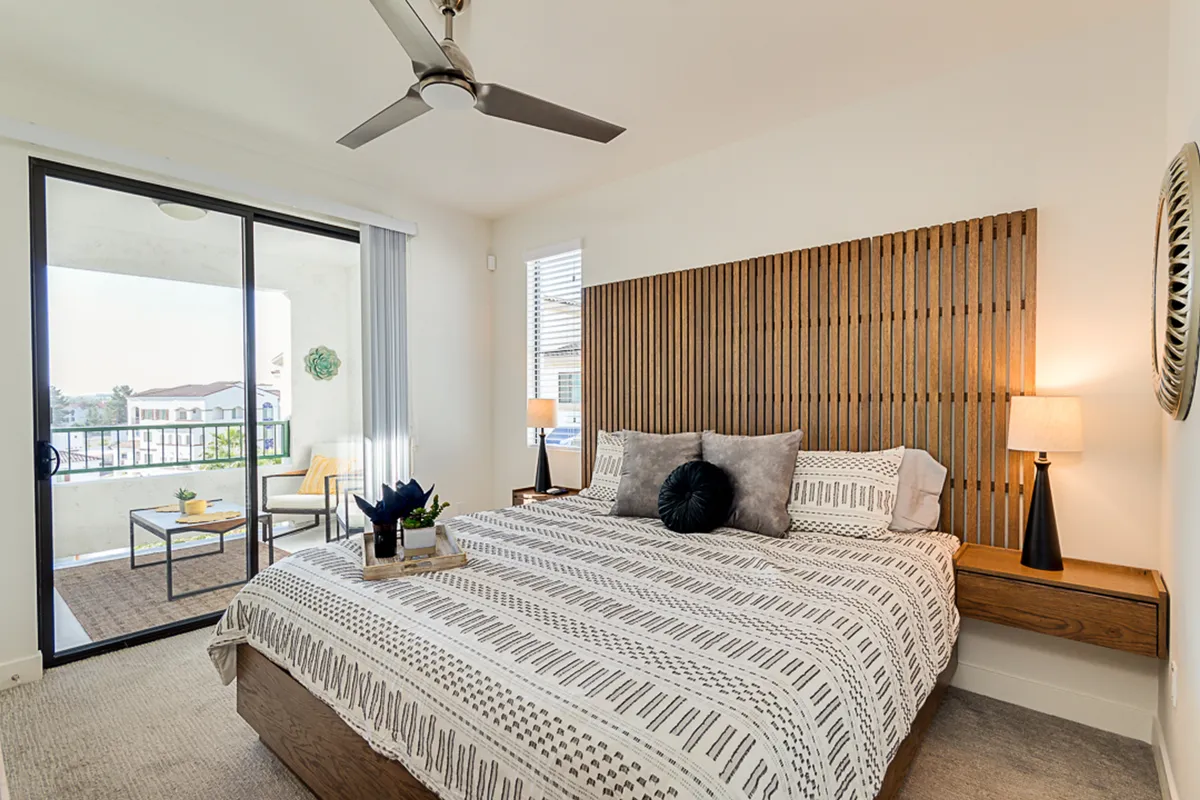 A serene bedroom scene with a bed featuring patterned bedding, a wooden headboard, table lamps, and a glimpse of balcony views beyond. The room is calm and inviting, with a clear connection to the outdoors through the balcony.