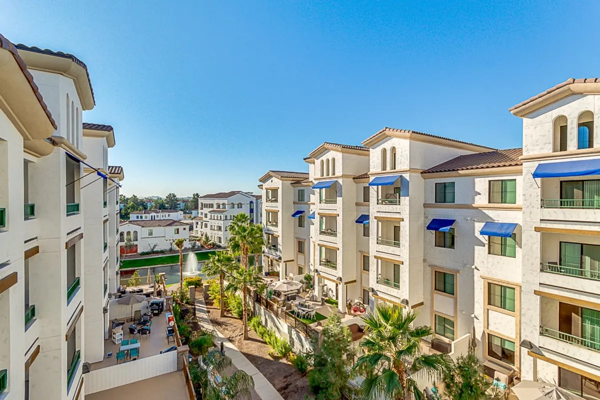 A welcoming exterior scene featuring balconies with distinctive blue awnings, surrounded by lush greenery and a tranquil fountain in the distance. The area is bathed in natural light, with visible pathways and gardens that invite leisurely strolls or moments of relaxation outdoors.