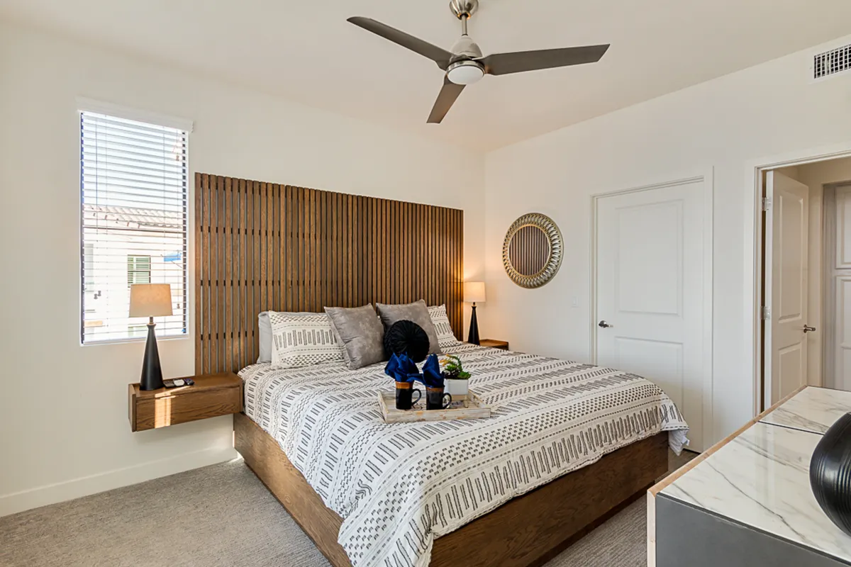 A cozy bedroom with a wooden headboard, bed adorned with decorative pillows, a table lamp, and a decorative mirror on the wall. Sunlight streams through the window, creating a warm and restful atmosphere ideal for relaxation and reading.