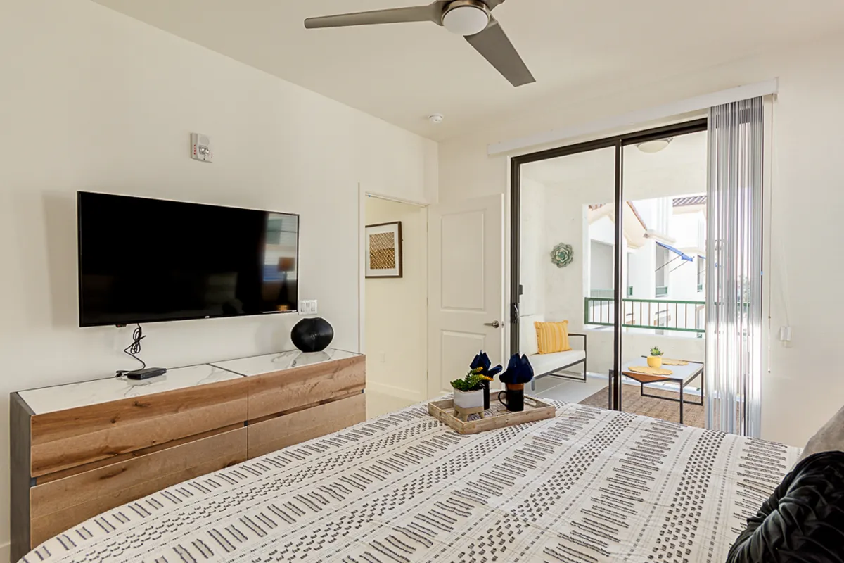 This image features a bed with patterned bedding, a flat-screen TV, a modern dresser, and a sliding glass door leading to a balcony. Natural light streams in from the balcony, hinting at an inviting view and easy access to outdoor relaxation.