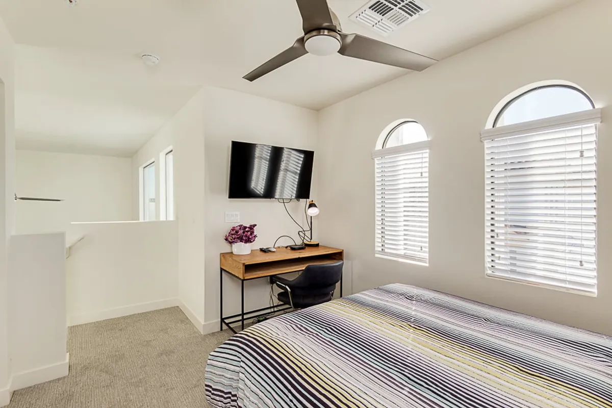 This image displays a bedroom with striped bedding, a dedicated desk with a lamp and chair ideal for working, a TV mounted on the wall for downtime, and arched windows with blinds that let in natural light. The workspace is integrated into the bedroom, embodying a seamless blend of productivity and comfort.